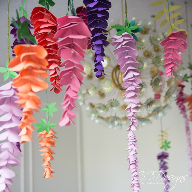 May include: A collection of paper wisteria flowers in various colours, including pink, purple, orange, and red, hanging from the ceiling. The flowers are made of paper and are arranged in clusters.