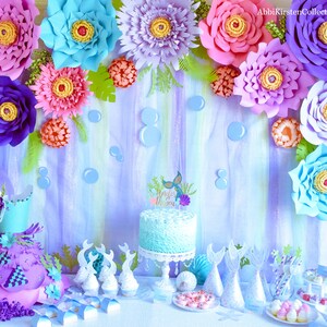 May include: A mermaid-themed party table setting with a blue and white cake, cupcakes, and other treats. The backdrop features paper flowers in shades of purple, pink, and blue, along with a painted ocean scene. The cake is decorated with a mermaid tail and the words "Little Mermaid".
