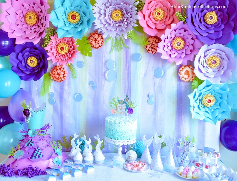 May include: A mermaid-themed party table setting with a blue and white cake, cupcakes, and other treats. The backdrop features paper flowers in shades of purple, pink, and blue, along with a painted ocean scene. The cake is decorated with a mermaid tail and the words "Little Mermaid".