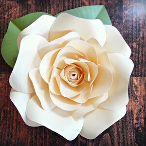 May include: A large, cream-colored paper flower with multiple layers of petals. The flower is on a brown wooden background.