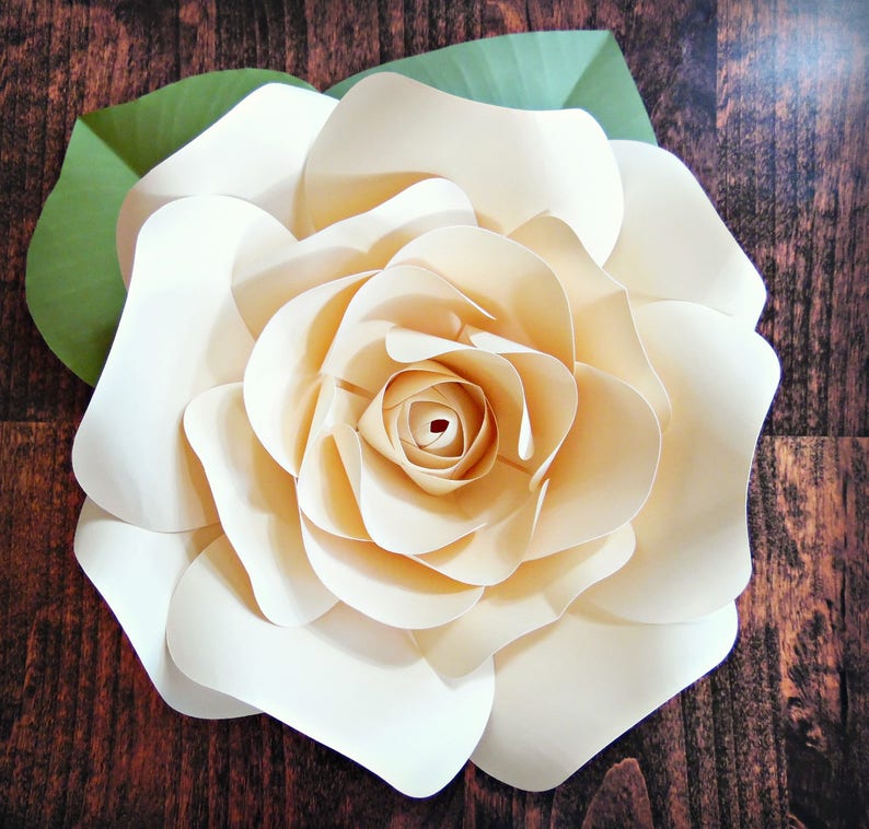 May include: A large, cream-colored paper flower with multiple layers of petals. The flower is on a brown wooden background.