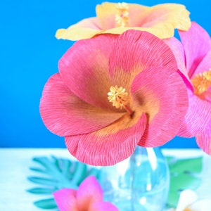 Puede incluir: Un ramo de flores de papel rosa y amarillo en un jarr&oacute;n de vidrio transparente. Las flores est&aacute;n dispuestas en un jarr&oacute;n sobre una superficie blanca con hojas verdes.