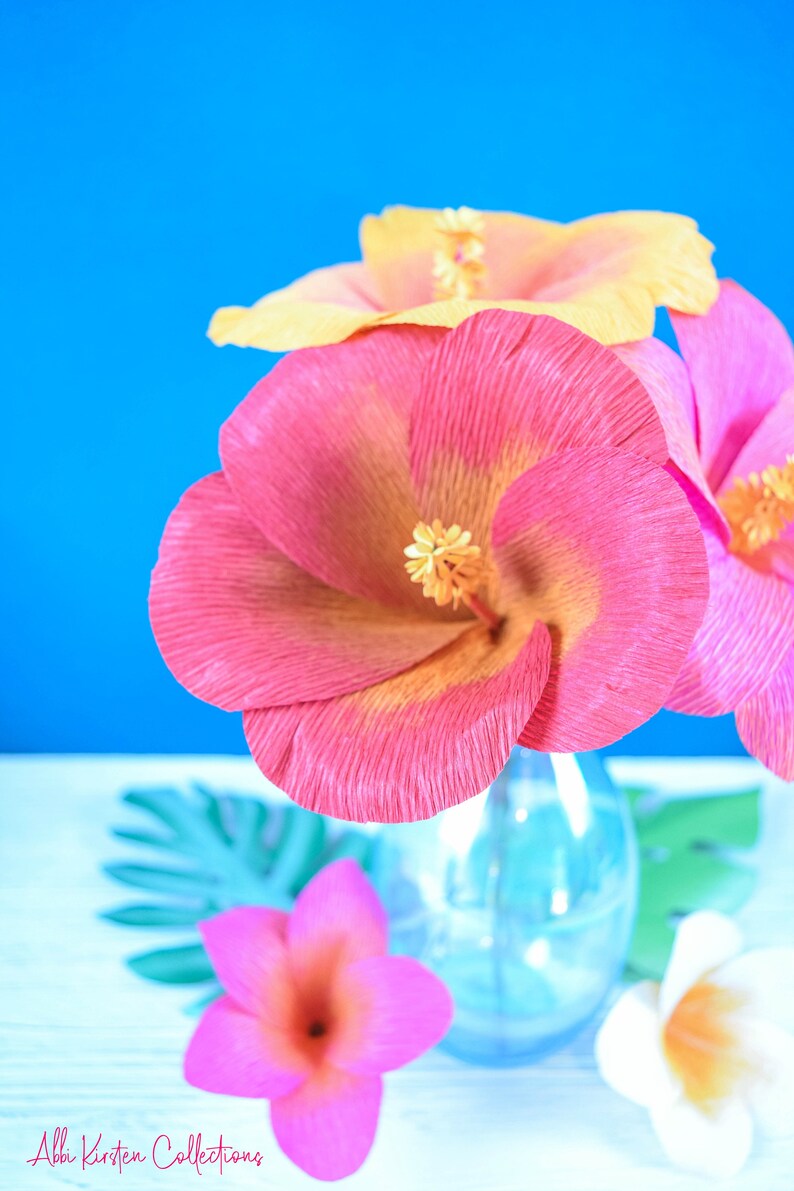 Puede incluir: Un ramo de flores de papel rosa y amarillo en un jarr&oacute;n de vidrio transparente. Las flores est&aacute;n dispuestas en un jarr&oacute;n sobre una superficie blanca con hojas verdes.