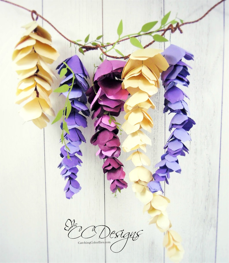 May include: A paper craft garland of wisteria flowers in shades of purple, lavender, and yellow. The flowers are arranged in clusters and hang from a brown twine string. The background is a white wood panel.