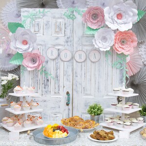 May include: A white, distressed door with a floral garland and the word "ROMA" in gray circles. A three-tiered white cupcake stand is filled with cupcakes. A table is set with a variety of food, including fruit, pastries, and cookies. The table is decorated with a white tablecloth and a silver metal tray.
