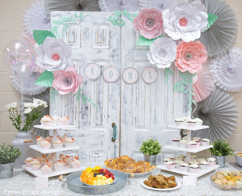 May include: A white, distressed door with a floral garland and the word "ROMA" in gray circles. A three-tiered white cupcake stand is filled with cupcakes. A table is set with a variety of food, including fruit, pastries, and cookies. The table is decorated with a white tablecloth and a silver metal tray.