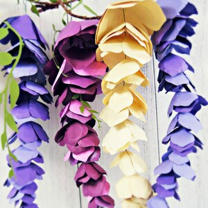 May include: Three paper wisteria flower garlands in shades of purple, lavender, and cream. The garlands are hanging from a branch and are made of individual paper flowers.