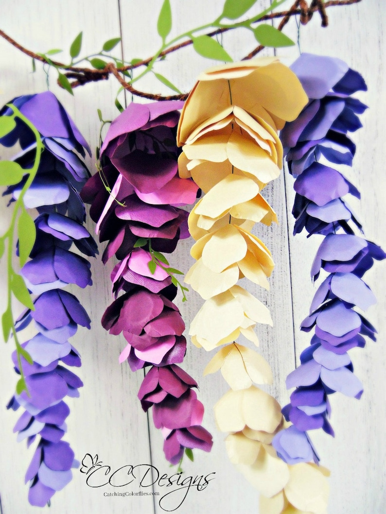 May include: Three paper wisteria flower garlands in shades of purple, lavender, and cream. The garlands are hanging from a branch and are made of individual paper flowers.