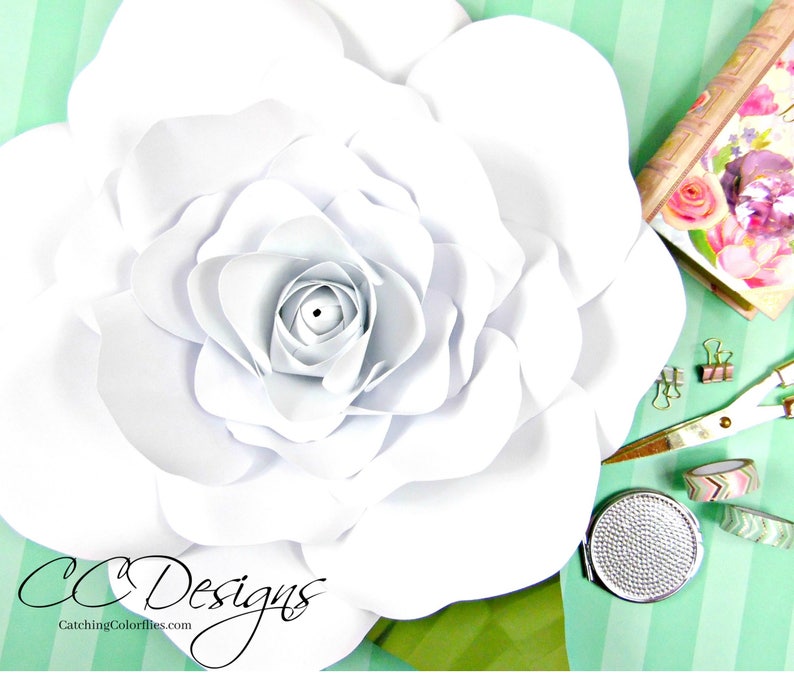May include: A large white paper flower with a layered center. The flower is on a green and white striped background. The text "CC Designs" and "CatchingColorflies.com" is visible in the bottom left corner of the image.
