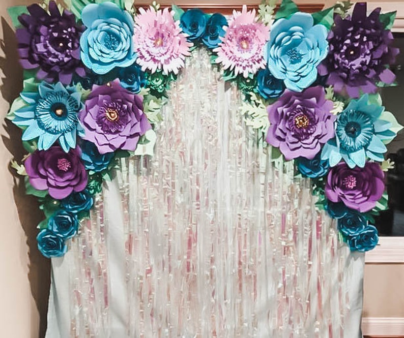 May include: A backdrop for a party or event, featuring a garland of large paper flowers in shades of purple, blue, and teal. The flowers are arranged in a semi-circle shape and are attached to a white backdrop with a shimmering, iridescent texture.