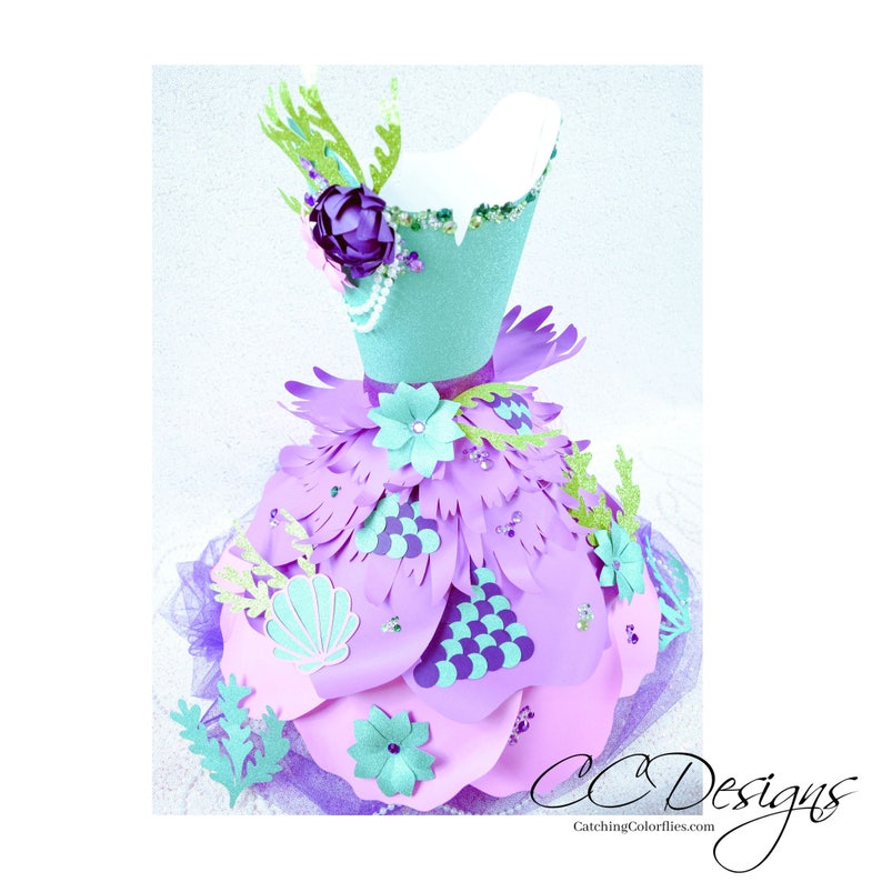 May include: A purple and teal papercraft dress with a mermaid theme. The dress is decorated with flowers, seashells, and scales. The dress is on a white mannequin.