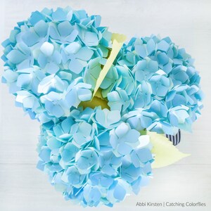 May include: A bouquet of light blue paper hydrangeas with yellow paper leaves. The hydrangeas are arranged in a vase with a striped pattern.