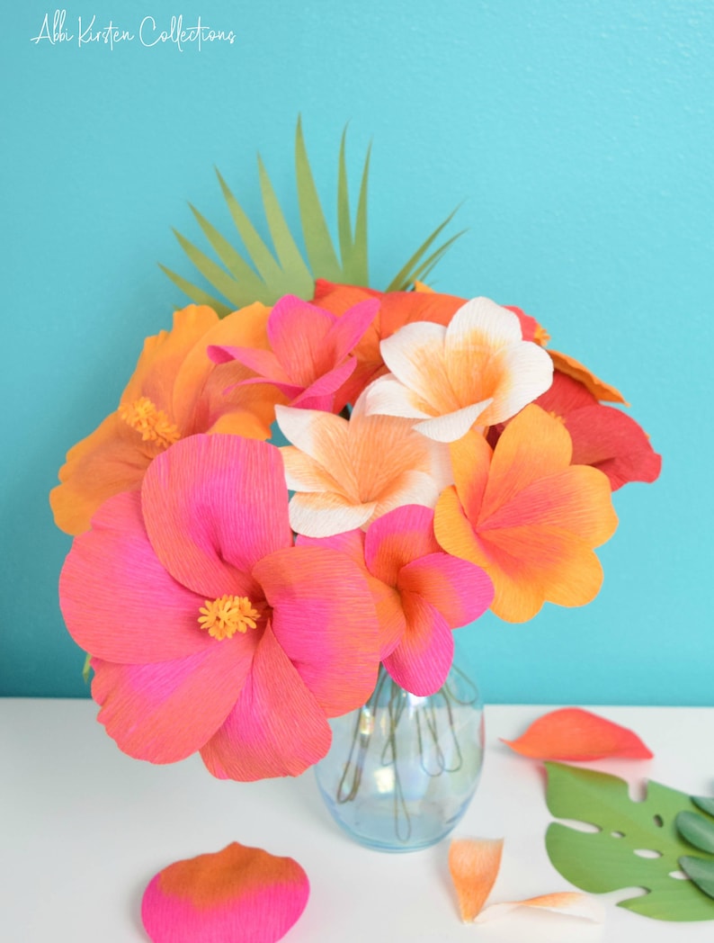 Puede incluir: Un ramo de flores de papel en un jarr&oacute;n de vidrio transparente. Las flores son rosas, naranjas y blancas y est&aacute;n dispuestas en un jarr&oacute;n sobre una superficie blanca. Tambi&eacute;n hay algunos p&eacute;talos sueltos en la superficie.