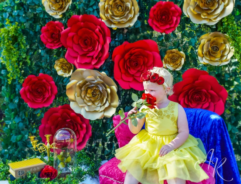 May include: A young girl wearing a yellow dress with a gold trim and a flower crown is smelling a red rose. She is sitting in front of a backdrop of red and gold paper roses.