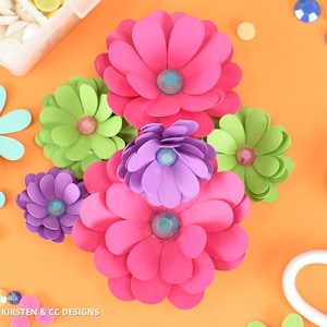 Puede incluir: Flores de papel coloridas en tonos rosa, morado y verde, con centros de gemas. Las flores están dispuestas sobre una superficie naranja, con suministros de manualidades como gemas y un recipiente con pequeños adornos blancos.