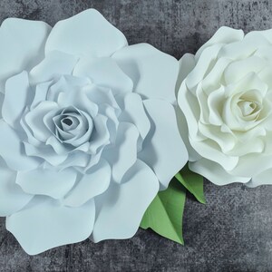 May include: Two large paper flowers, one light blue and one white, with green leaves. The flowers are made of paper and have a layered, ruffled appearance.