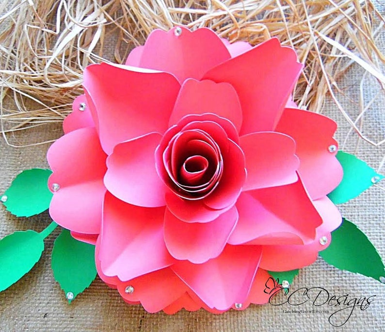 May include: A large, pink paper rose with a green leaf stem. The rose is made of multiple layers of paper and has a center that is curled inward. The rose is decorated with small, clear rhinestones.