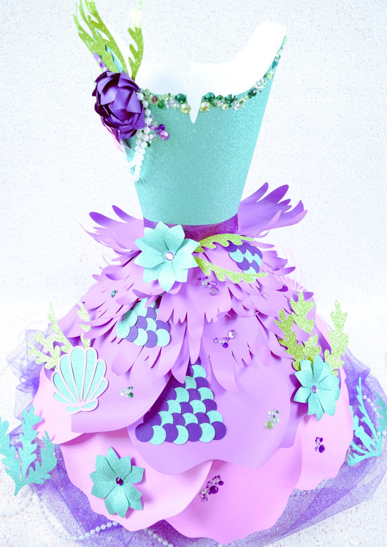 May include: A papercraft mermaid dress with a turquoise bodice, purple flowers, and a pink skirt with green seaweed and seashells. The skirt is made of layers of paper and has a tulle overlay.