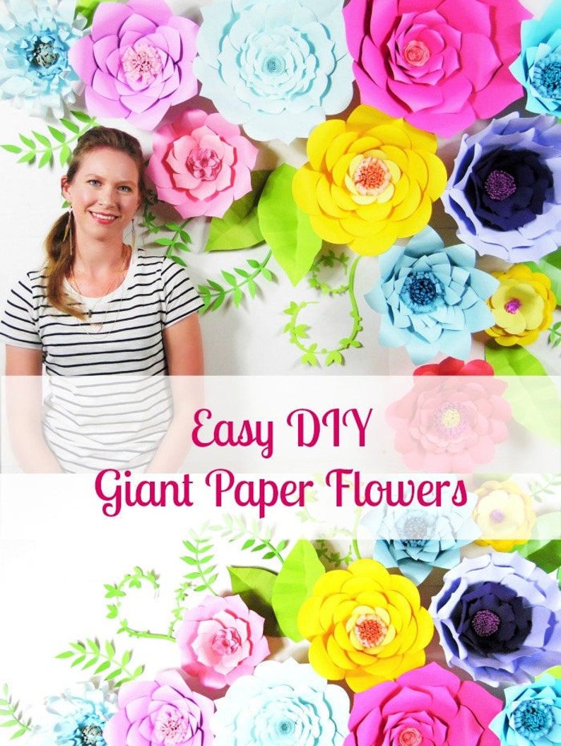 May include: A woman stands in front of a wall decorated with large, colorful paper flowers. The flowers are in various shades of pink, blue, yellow, and purple. The text "Easy DIY Giant Paper Flowers" is written in pink letters across the bottom of the image.