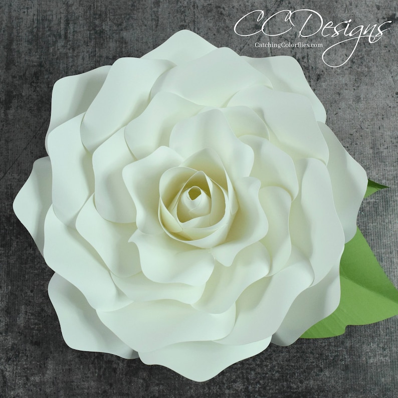May include: A large, white paper flower with multiple layers of petals. The flower is made to look like a rose and has a green leaf attached to the side.