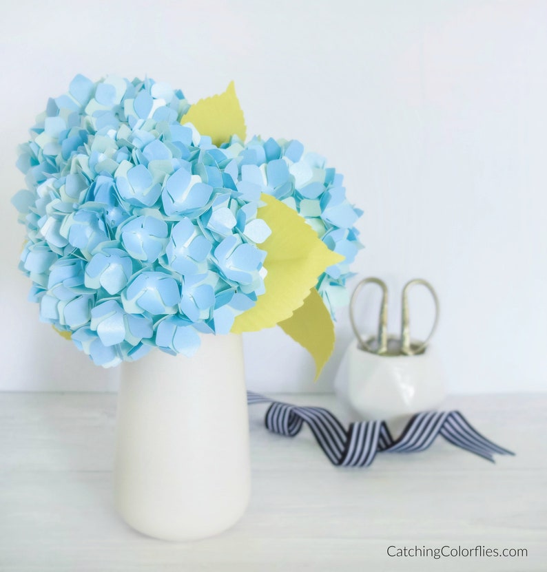 May include: A paper flower arrangement in a white vase. The flowers are blue and white hydrangeas with yellow leaves. The vase is on a white wooden table.