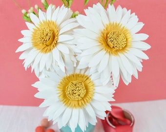 Op de afbeelding: Drie witte papieren margrieten met gele centra in een turquoise vaas. De margrieten zijn in een boeket gerangschikt en vormen het middelpunt van de afbeelding.