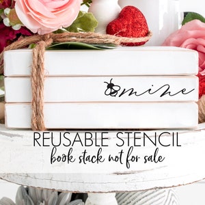 May include: A white book stack with the words "bee mine" written on the top book. The book stack is decorated with twine and is surrounded by pink and red flowers. The book stack is on a white tiered tray with wooden beads.