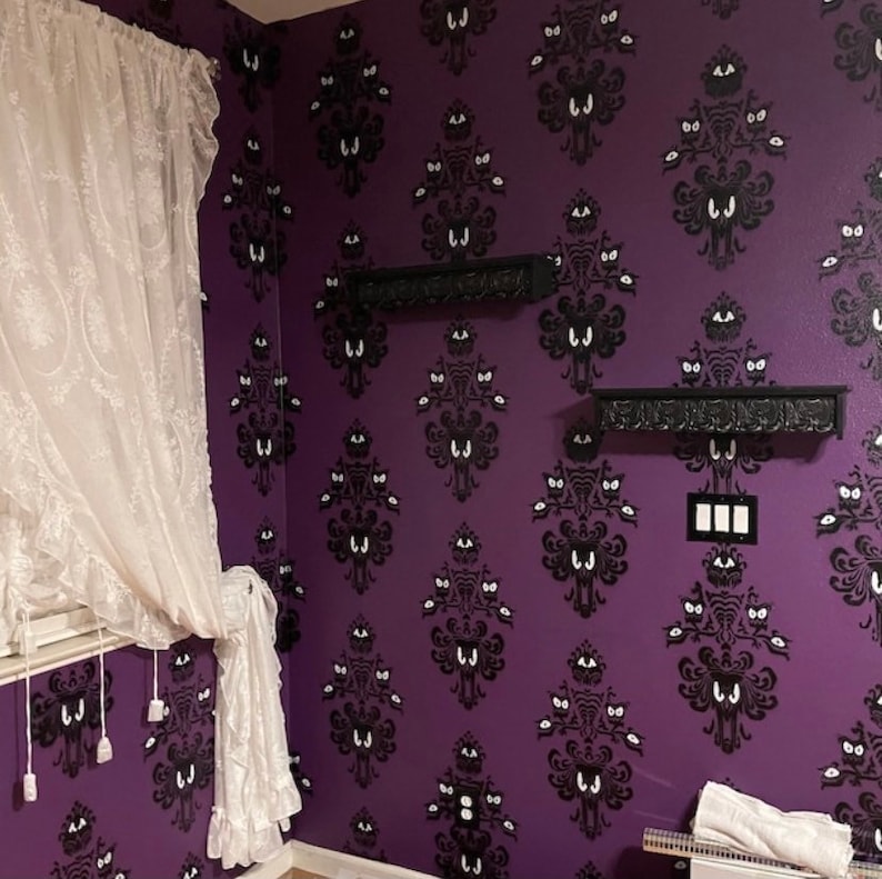 May include: Purple wall with a black and white wallpaper pattern featuring a repeating design of a ghost with a single eye. Two black shelves are mounted on the wall. A white lace curtain is on the left side of the image.