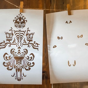 May include: Two white stencils for painting. The first stencil features a detailed design of a haunted house with a spooky, gothic style. The second stencil features a set of eyes, a nose, and a mouth.
