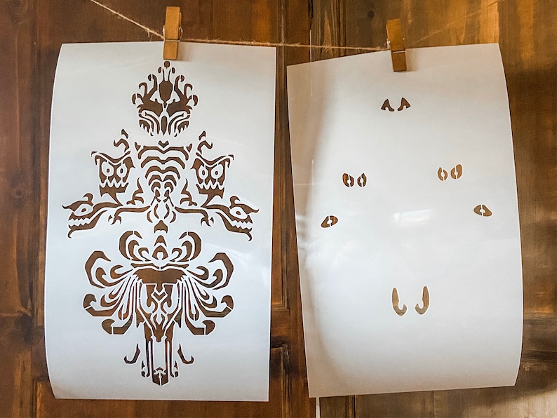 May include: Two white stencils for painting. The first stencil features a detailed design of a haunted house with a spooky, gothic style. The second stencil features a set of eyes, a nose, and a mouth.
