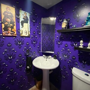 May include: A purple bathroom with a white porcelain sink and toilet. The walls are covered in a wallpaper pattern of ghostly faces. There are two framed pictures on the wall, one of a man in a top hat and the other of a woman in a dress. There is a mirror above the sink and a shelf above the toilet.