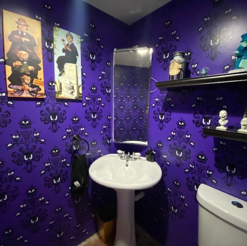May include: A purple bathroom with a white porcelain sink and toilet. The walls are covered in a wallpaper pattern of ghostly faces. There are two framed pictures on the wall, one of a man in a top hat and the other of a woman in a dress. There is a mirror above the sink and a shelf above the toilet.
