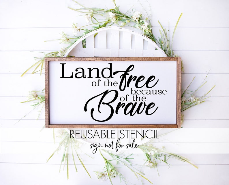Pode incluir: Uma placa de madeira com o texto "Land of the free because of the brave" em letras pretas. A placa &eacute; enquadrada em uma moldura de madeira marrom e tem um fundo branco. A placa &eacute; cercada por flores brancas e folhas verdes. O texto "Reusable Stencil sign not for sale" &eacute; impresso abaixo da placa.