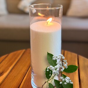 May include: A white pillar candle with a flame burning inside a clear glass jar. The candle is on a wooden table with a sprig of white flowers.