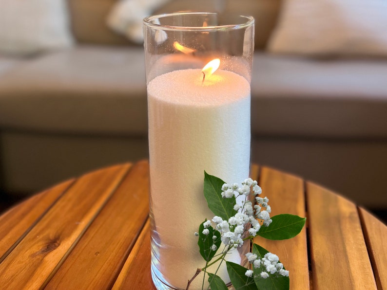 May include: A white pillar candle with a flame burning inside a clear glass jar. The candle is on a wooden table with a sprig of white flowers.