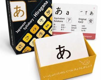 May include: A Japanese Hiragana flashcard set by Cardoia featuring a black storage box with yellow character tiles, a white instructional card displaying the character 'a' with its Katakana and Kanji equivalents, and a yellow box containing white cards.