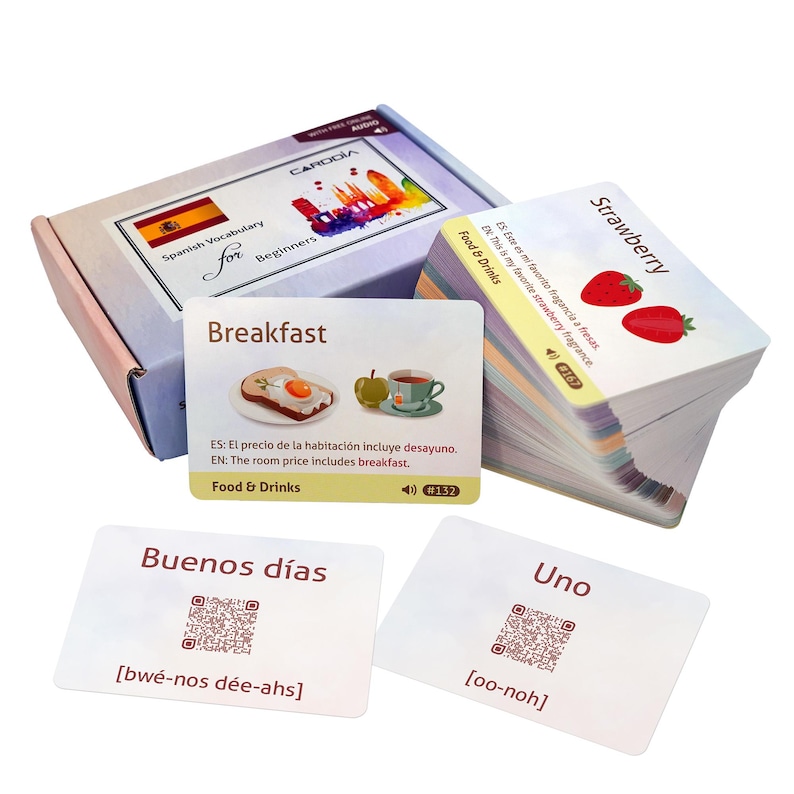 May include: A set of Spanish vocabulary flashcards for beginners, including a storage box, a stack of cards, and individual cards featuring words like Breakfast, Strawberry, Buenos d&iacute;as, and Uno, with illustrations, QR codes, and English translations.