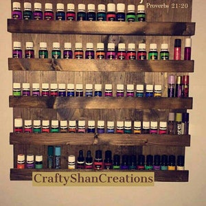May include: A wooden shelf unit with four shelves, each holding rows of small glass bottles with white lids. The bottles are filled with colorful liquids. The top shelf has a sign that reads "There is oil in the house of the wise Proverbs 21:20". The bottom shelf has the text "CraftyShanCreations".