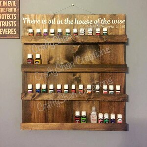 May include: A wooden wall shelf with three shelves for displaying essential oil bottles. The shelf is stained a dark brown color and has a white inscription that reads "There is oil in the house of the wise Proverbs 21:20".
