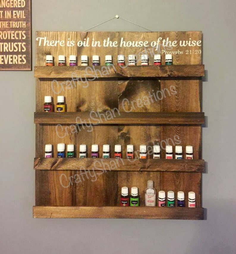 May include: A wooden wall shelf with three shelves for displaying essential oil bottles. The shelf is stained a dark brown color and has a white inscription that reads "There is oil in the house of the wise Proverbs 21:20".
