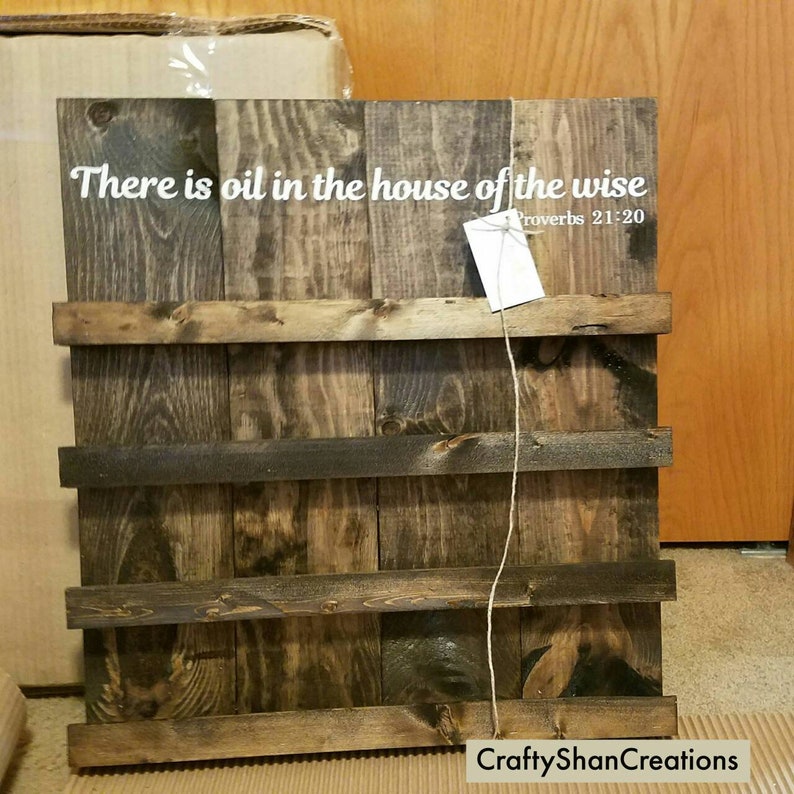 May include: A rustic wooden sign with the text "There is oil in the house of the wise" and the scripture reference "Proverbs 21:20". The sign has a dark brown stain and is made of reclaimed wood.