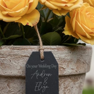 May include: A bouquet of vibrant yellow roses in a textured, weathered beige terracotta pot. A rectangular black slate tag hangs from the roses by a jute twine string, engraved with the text: On your Wedding Day, Andrew & Eliza, With Love, Nicola & Simon, 5.7.2025.