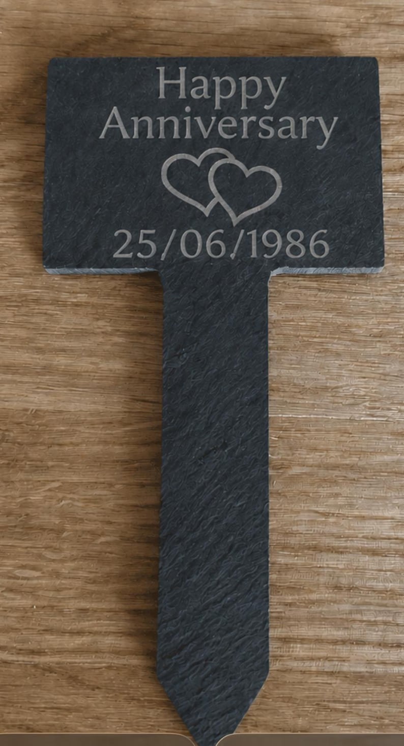 May include: A dark gray slate garden marker with a rectangular top and a pointed stake base. The surface is engraved with the text Happy Anniversary, a graphic of two overlapping hearts, and the date 25/06/1986 in a light gray font.