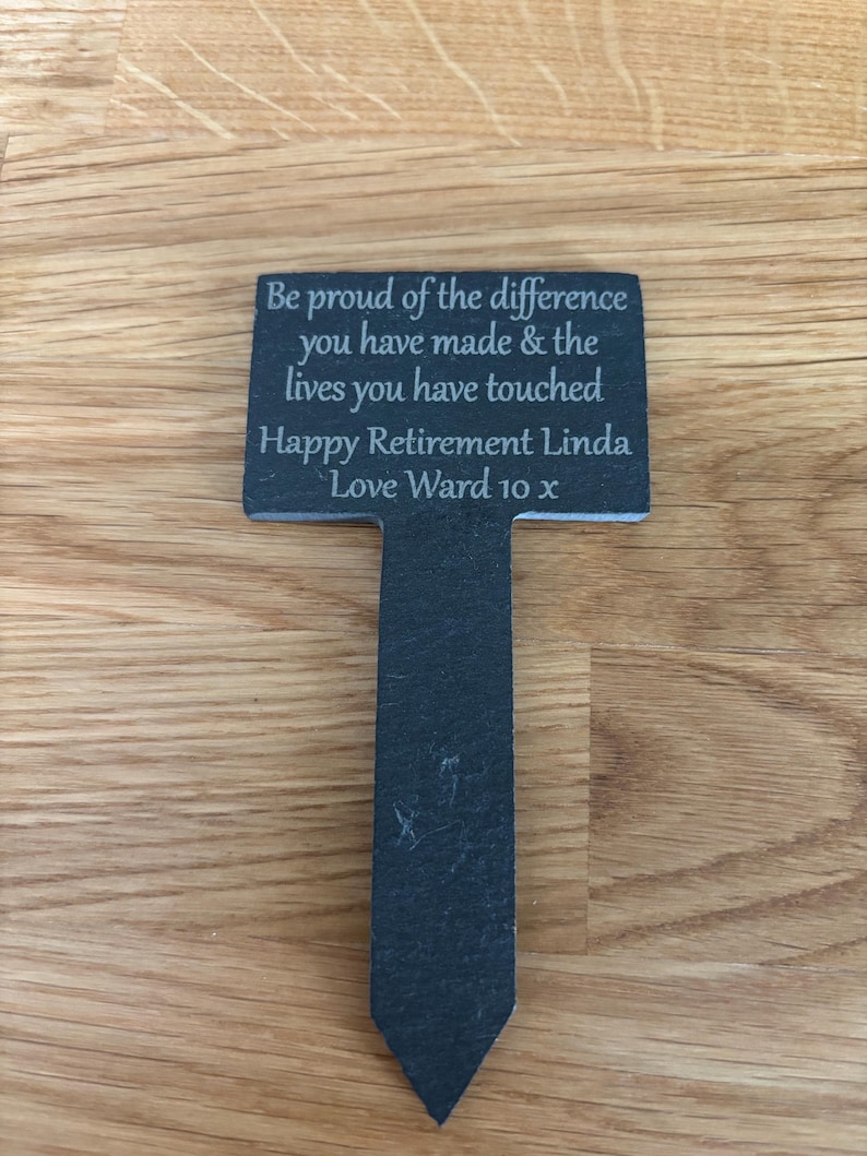 May include: A T-shaped slate garden marker with engraved text that reads: Be proud of the difference you have made & the lives you have touched. Happy Retirement Linda. Love Ward 10 x. The dark gray stone has a natural texture and a pointed bottom stake.
