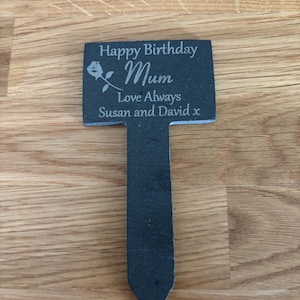 May include: A dark gray slate garden marker with a pointed stake base. The rectangular top features an engraved rose graphic and the text: Happy Birthday Mum, Love Always, Susan and David x. The surface has a natural, textured stone finish.