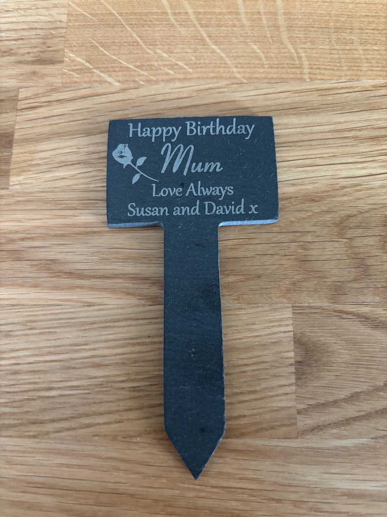 May include: A dark gray slate garden marker with a pointed stake base. The rectangular top features an engraved rose graphic and the text: Happy Birthday Mum, Love Always, Susan and David x. The surface has a natural, textured stone finish.