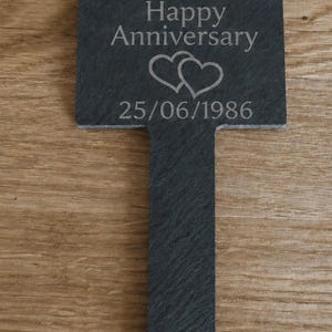 May include: A dark gray slate garden marker with a rectangular top and a pointed stake base. The surface is engraved with the text Happy Anniversary, two overlapping heart outlines, and the date 25/06/1986 in a light gray sans-serif font.