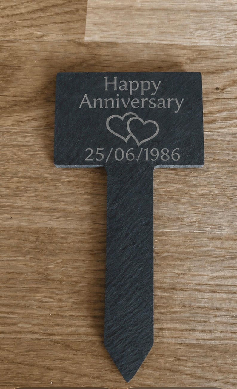 May include: A dark gray slate garden marker with a rectangular top and a pointed stake base. The surface is engraved with the text Happy Anniversary, two overlapping heart outlines, and the date 25/06/1986 in a light gray sans-serif font.