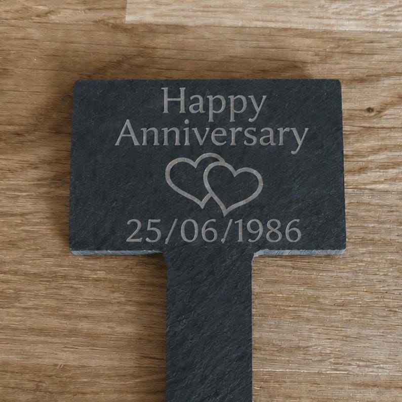 May include: A rectangular dark gray slate garden marker featuring the engraved text Happy Anniversary, a graphic of two overlapping hearts, and the date 25/06/1986. The marker has a vertical stake extending from the bottom center of the slate plaque.