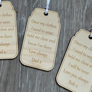May include: Three wooden tags shaped like mason jars with a white ribbon attached. The tags have a sentimental message engraved on them. The first tag says "Once my clothes I used to wear, hold me close and know I'm there Love always Dad x". The second tag says "Once my clothes I used to wear, hold me close and know I'm there Love always Dad x". The third tag says "Once my clothes I used to wear, hold me close and I will be near Love you always Bob x".